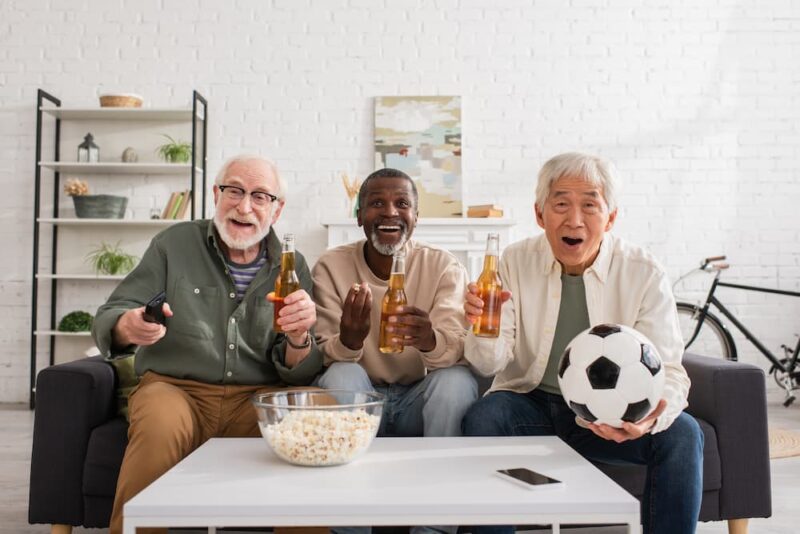 Three people sitting on a couch, enjoying beers and popcorn. One holds a soccer ball. They're in a cozy living room setting.