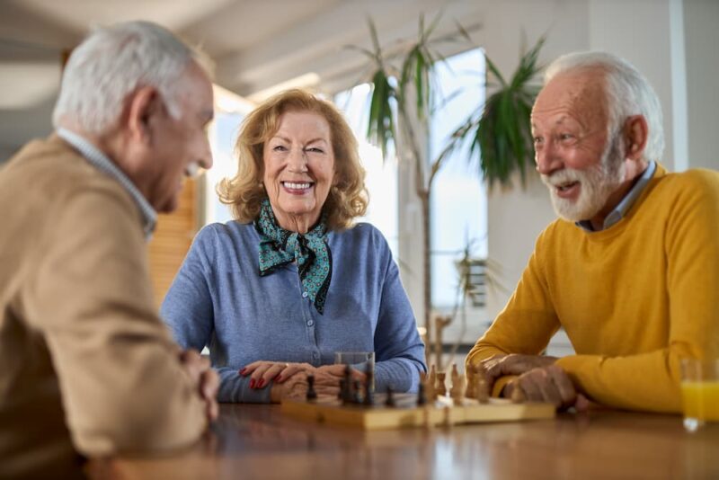 Three people seated at a table, smiling and playing chess, with a plant in the background. Bright, cheerful indoor setting.