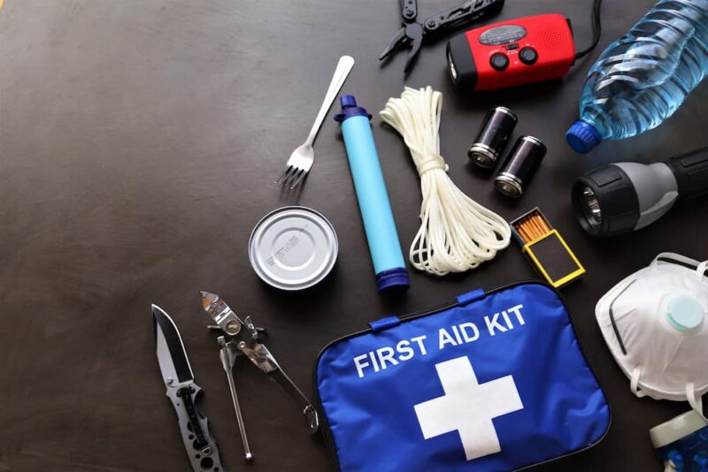 Emergency supplies, including a first aid kit, flashlight, rope, knife, canned food, water, radio, LifeStraw, and matches, are arranged on a dark surface.