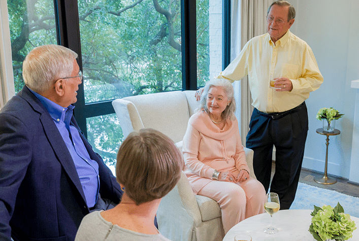 Four people are gathered indoors, conversing and holding drinks. A person is seated, while three others stand around. Large windows reveal trees outside.