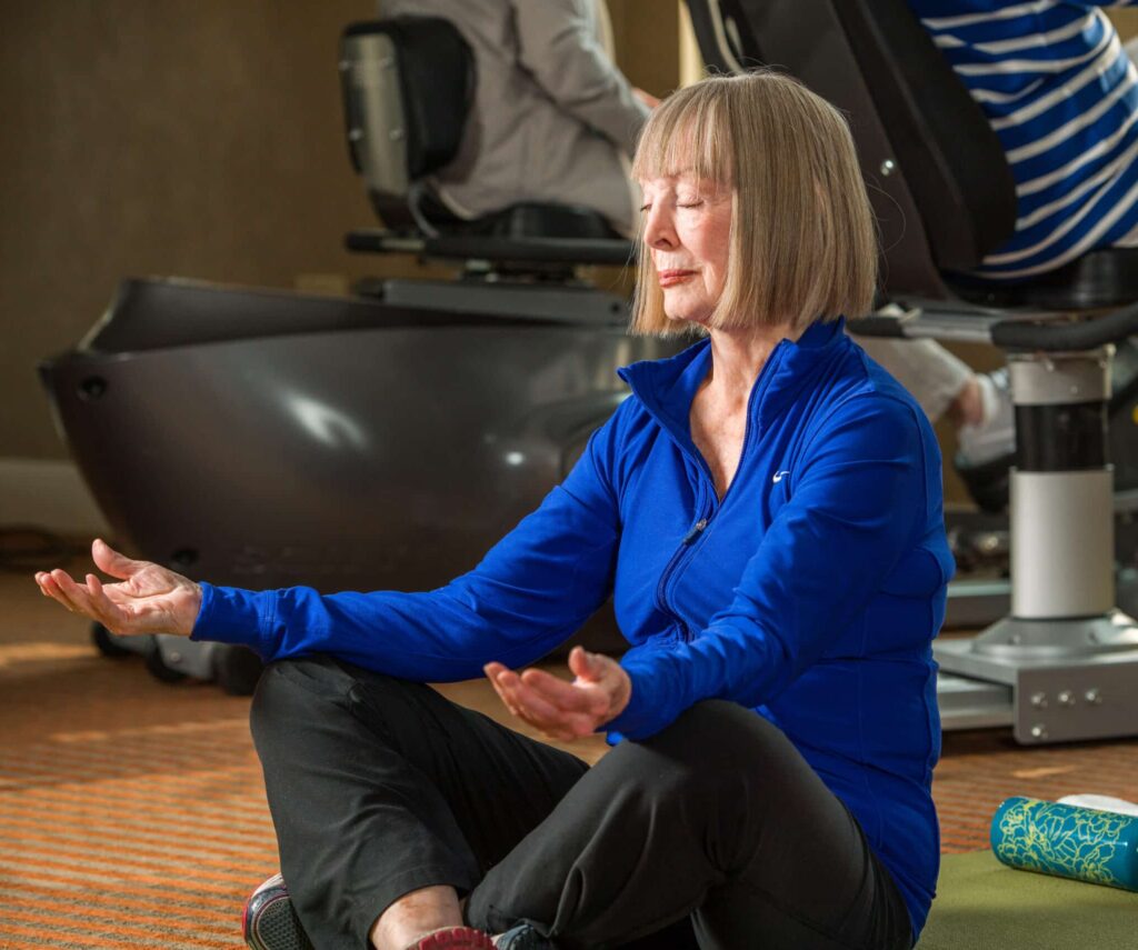 Elderly woman in blue jumper and black athletic pants sits crossed-legged while meditating in a yoga pose.
