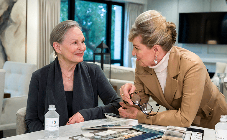 Two people interact at a table with makeup palettes and bottled water. One sits, smiling, while the other leans forward, holding glasses.