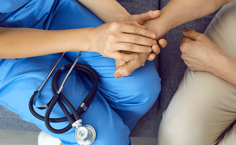 A person in blue scrubs, with a stethoscope, comforting another person by holding their hand and sitting together on a couch.