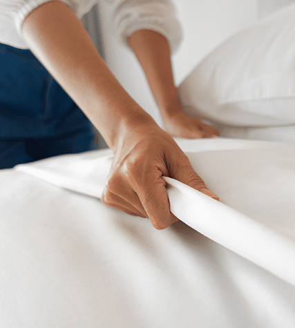 A person neatly tucks in white bed sheets, creating smooth lines. The focus is on their hands, showcasing careful and precise bed-making.