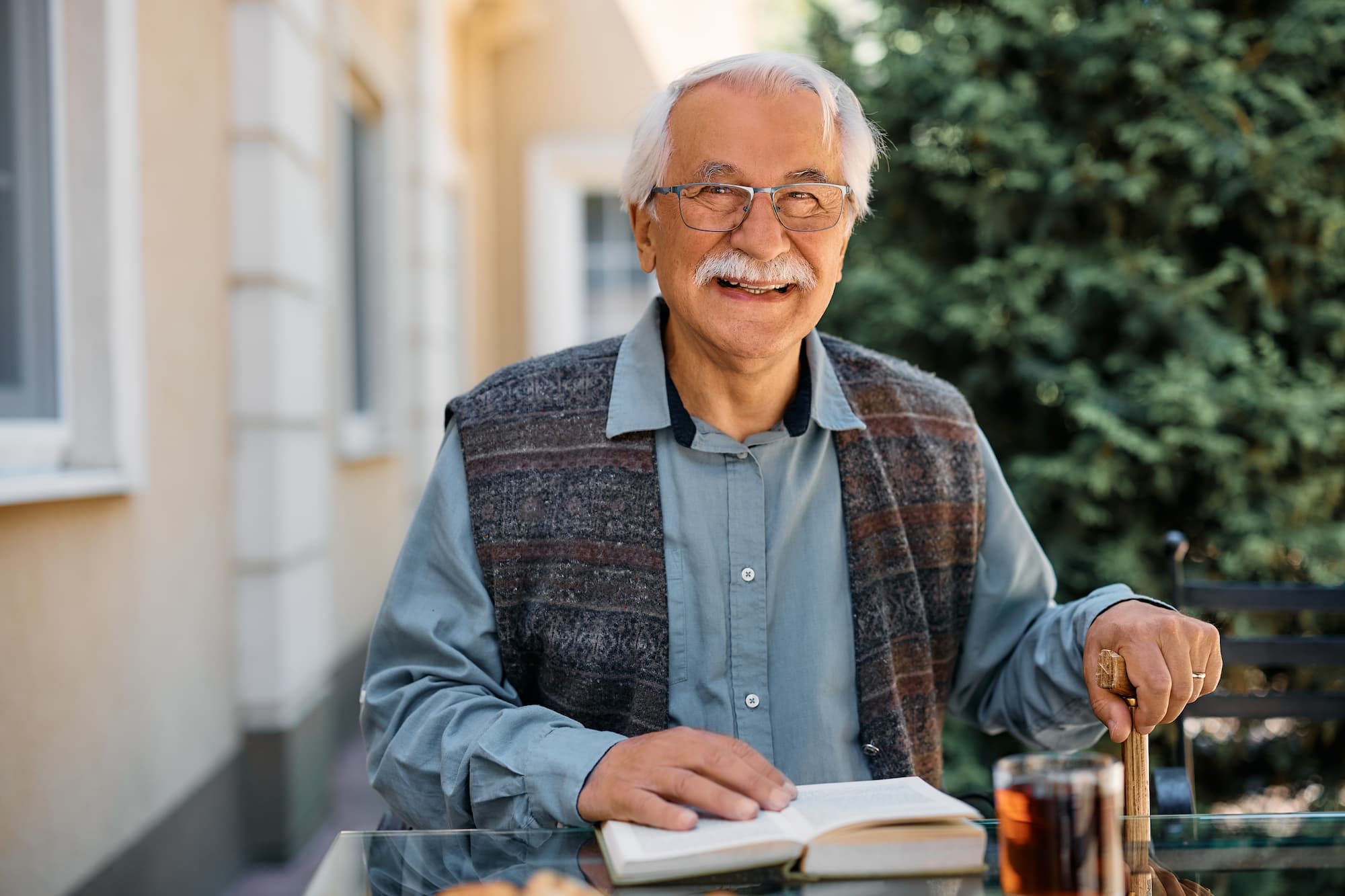 A smiling person with glasses, sitting outdoors at a table, reading a book. There's a glass of tea and greenery in the background.