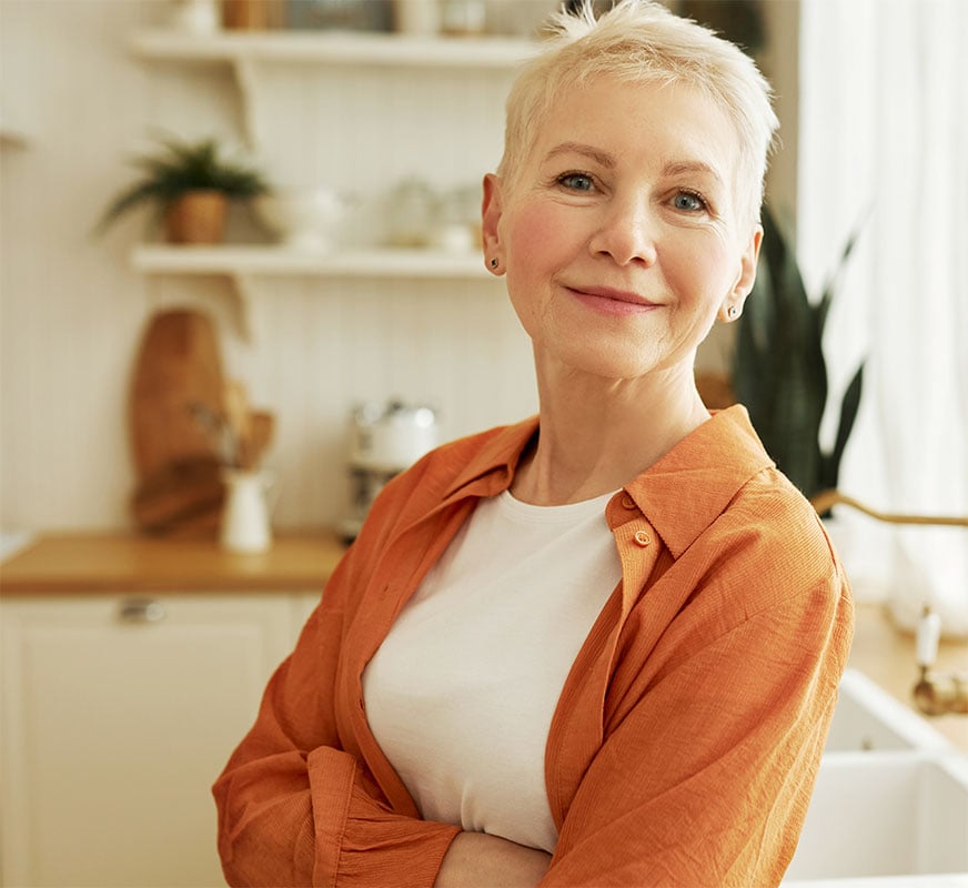 A woman with short hair, wearing a white shirt and orange jacket, smiles in a cozy kitchen with white shelves and houseplants.